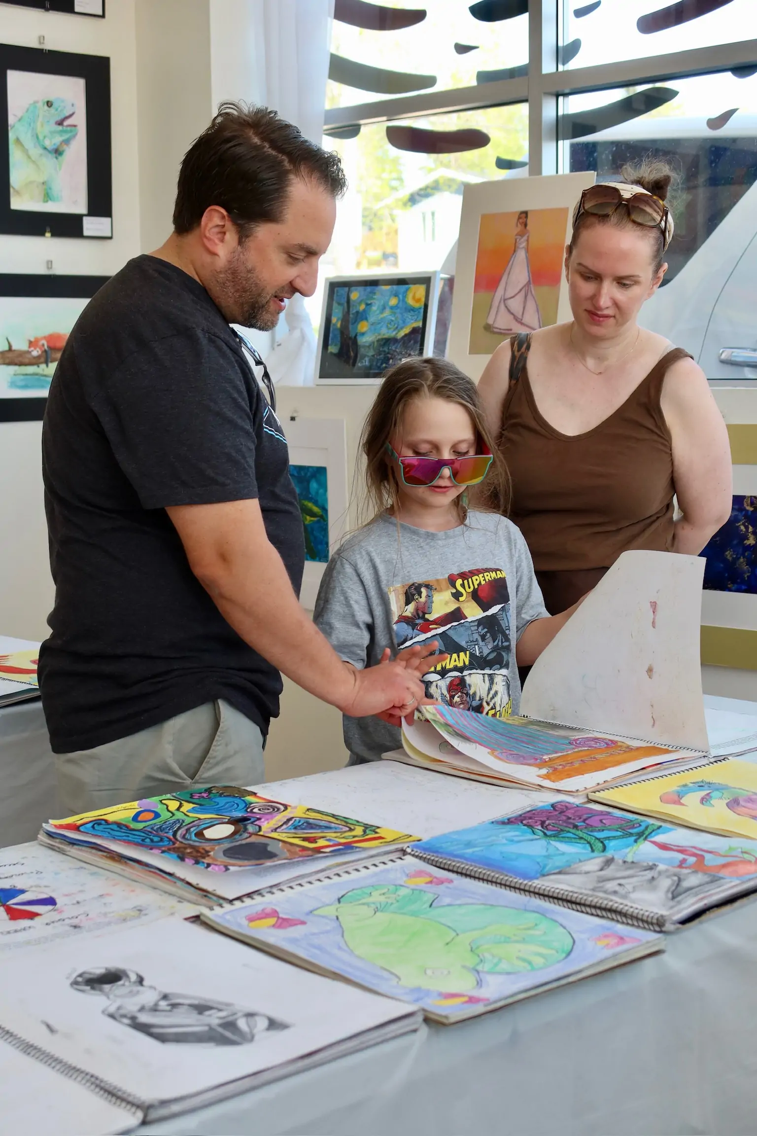 A child looks through their artwork while standing with two adults during the student art showcase at Aliki’s Art House.